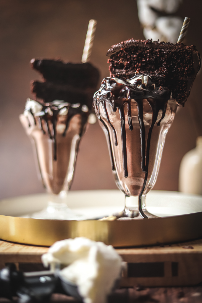 A photo of two chocolate cake milkshakes with a slice of chocolate cake on top