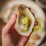 A photo of someone holding an oyster on the half shell with tomatillo habanero mignonette