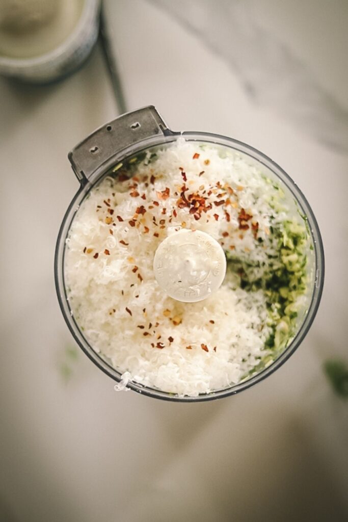 A food processor with almonds, arugula parmesan cheese and chili flakes.