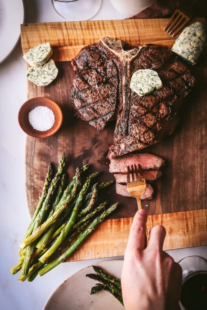 A grilled t-bone steak with asparagus and someone taking a few slices.