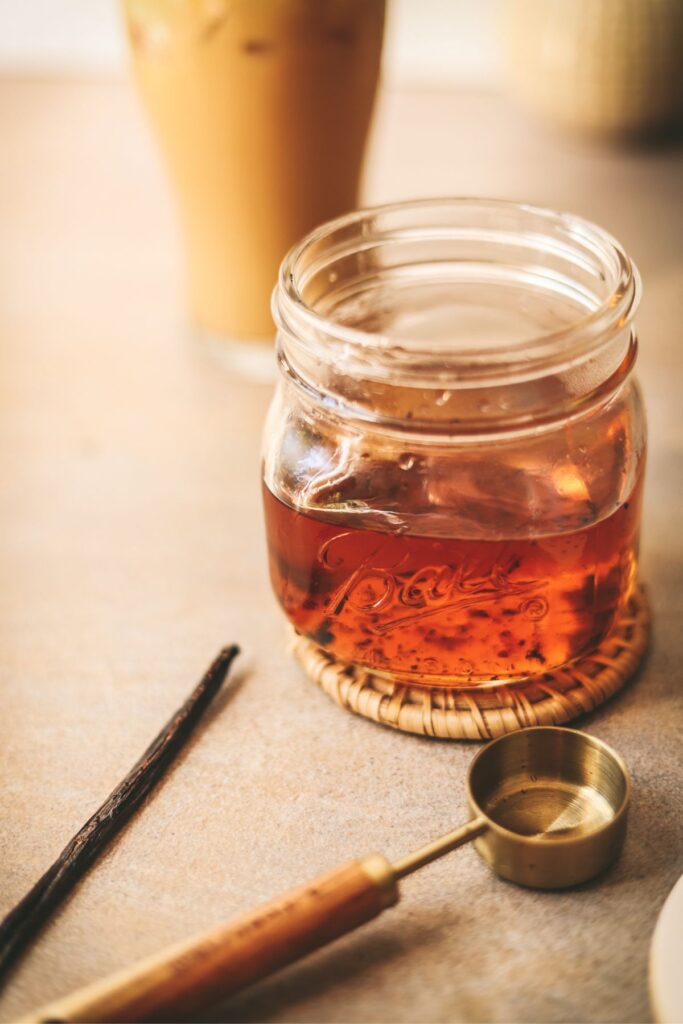 A jar of vanilla syrup with a vanilla bean and wooden measuring spoon. 