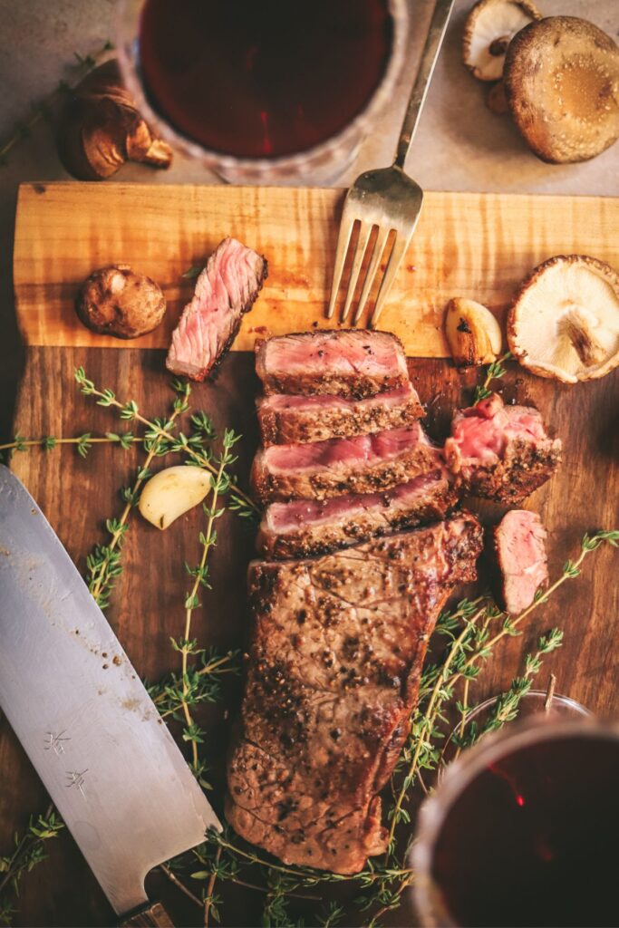 A steak on a wooden cutting board with thyme, mushrooms, a knife and red wine. 