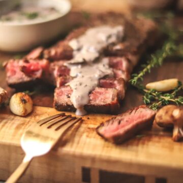Steak covered in mushroom sauce on a wooden cutting board with herbs.