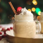 Two eggnog lattes in front of a Christmas tree with whipped cream.