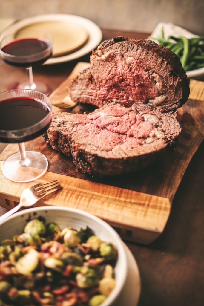 A prime rib meal on a cutting board with red wine and sides. 