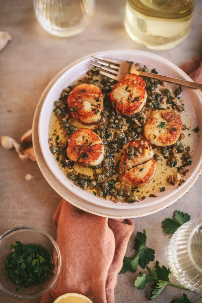 Five seared scallops in lemon caper butter, golden brown, with a fork and white wine. 