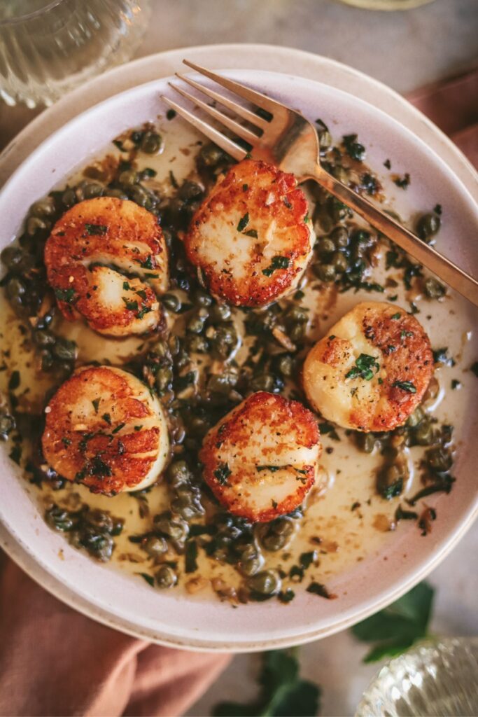 Five seared scallops on a plate with lemon and caper butter. 