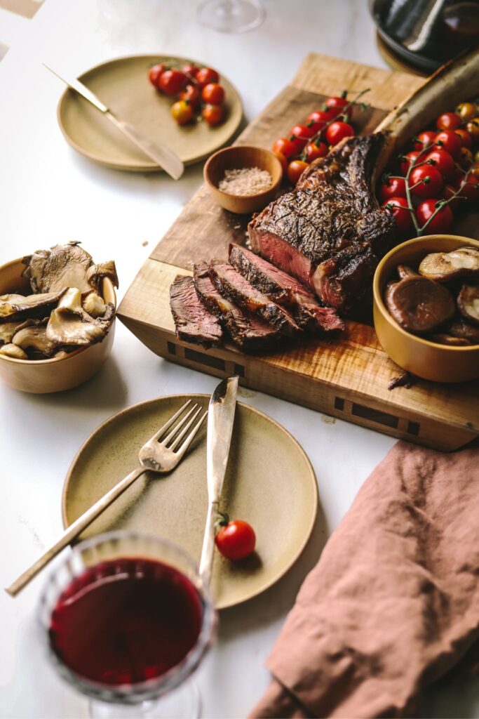 A tomahawk steak on a wooden cutting board with red wine, mushrooms and tomatoes. 