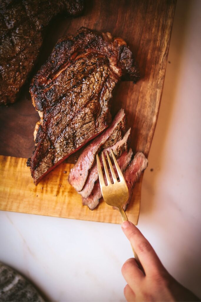 Grilled ribeye steak on a wooden cutting board, sliced, with someone grabbing a bite with a fork. 