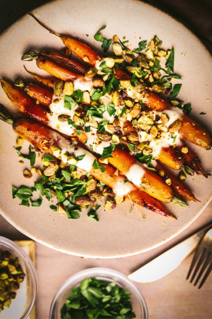 A plate of finished roasted carrots tossed in a honey glaze. garnished with orange creme fraiche, pistachios, and parsley. 