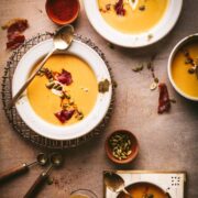 Several bowls of roasted butternut squash soup, surrounded by garnishes like pumpkin seeds and chili powder.