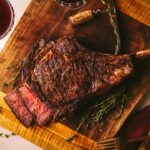 A perfectly seared tomahawk steak, cooked medium rare, on a wooden cutting board with red wine.