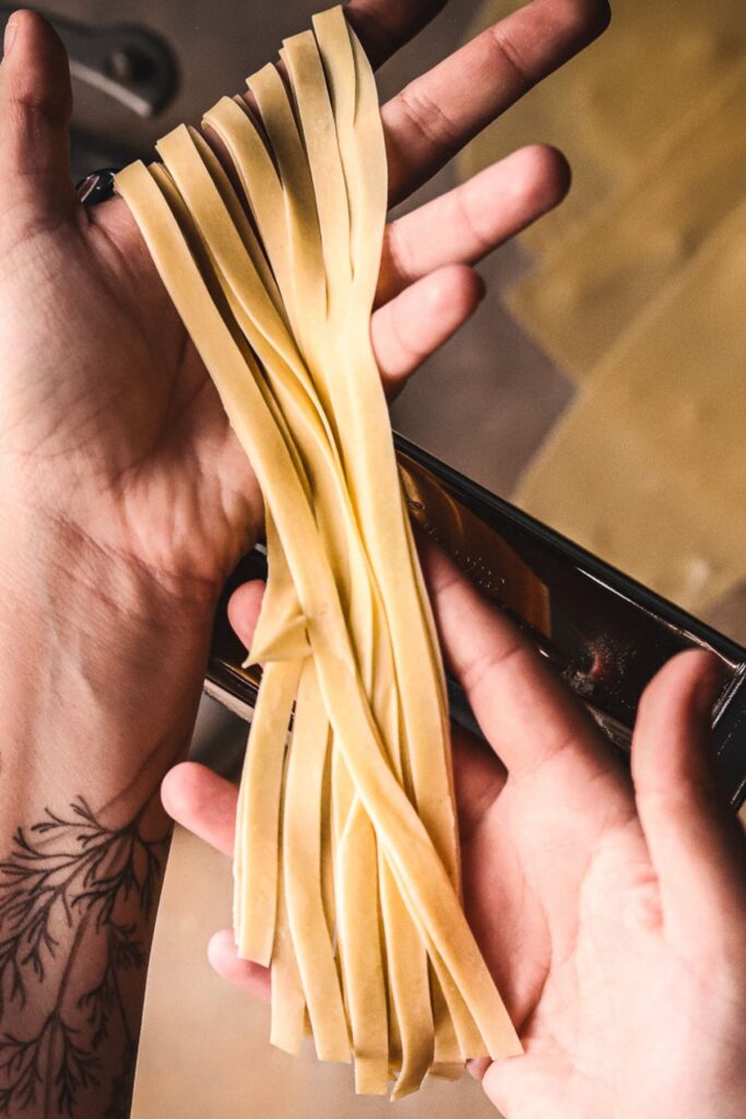 Two hands holding freshly cut homemade fettucine over a pasta roller. 
