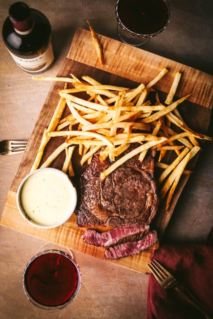 A wooden cutting board with steak, homemade French fries (frites), Béarnaise sauce, and red wine. 