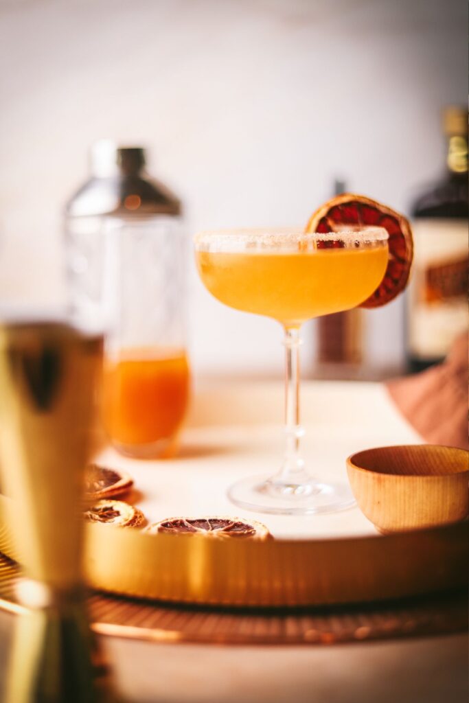 A photo of an apple sidecar cocktail, with dried citrus, a jigger, and cider. 